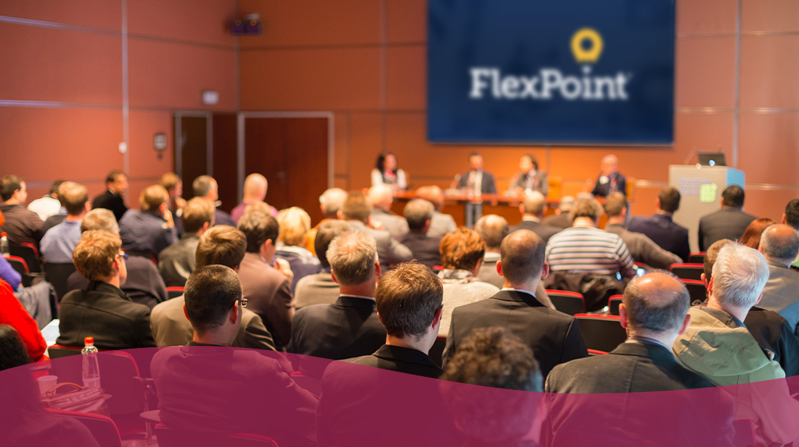 An audience seated facing a stage with a panel of speakers and a screen displaying