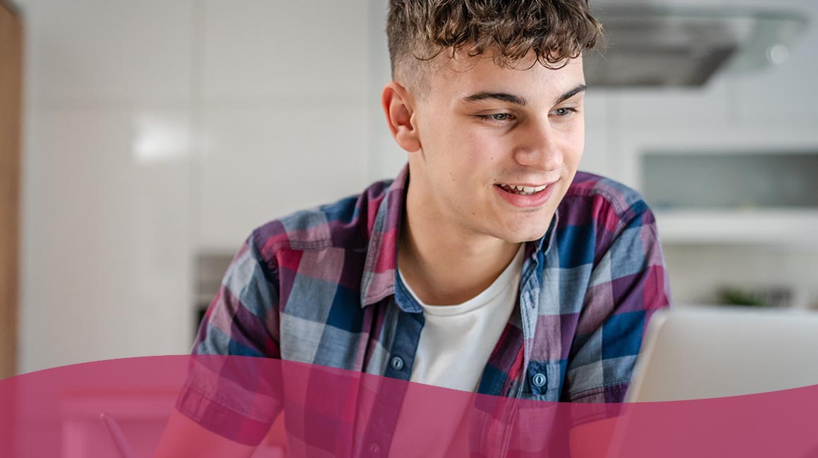 A young person attending a virtual school smiling at a laptop, wearing a blue and pink plaid shirt in a kitchen setting.
