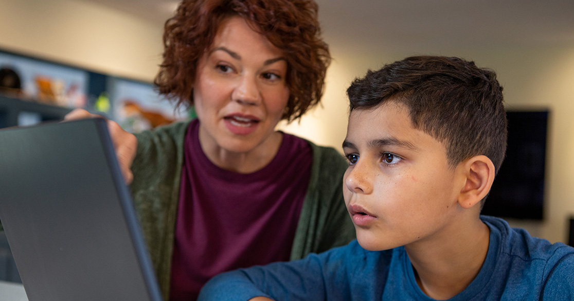 A child works on a laptop at a table while an adult provides guidance, illustrating a home-based digital learning environment with a notebook nearby for taking notes.